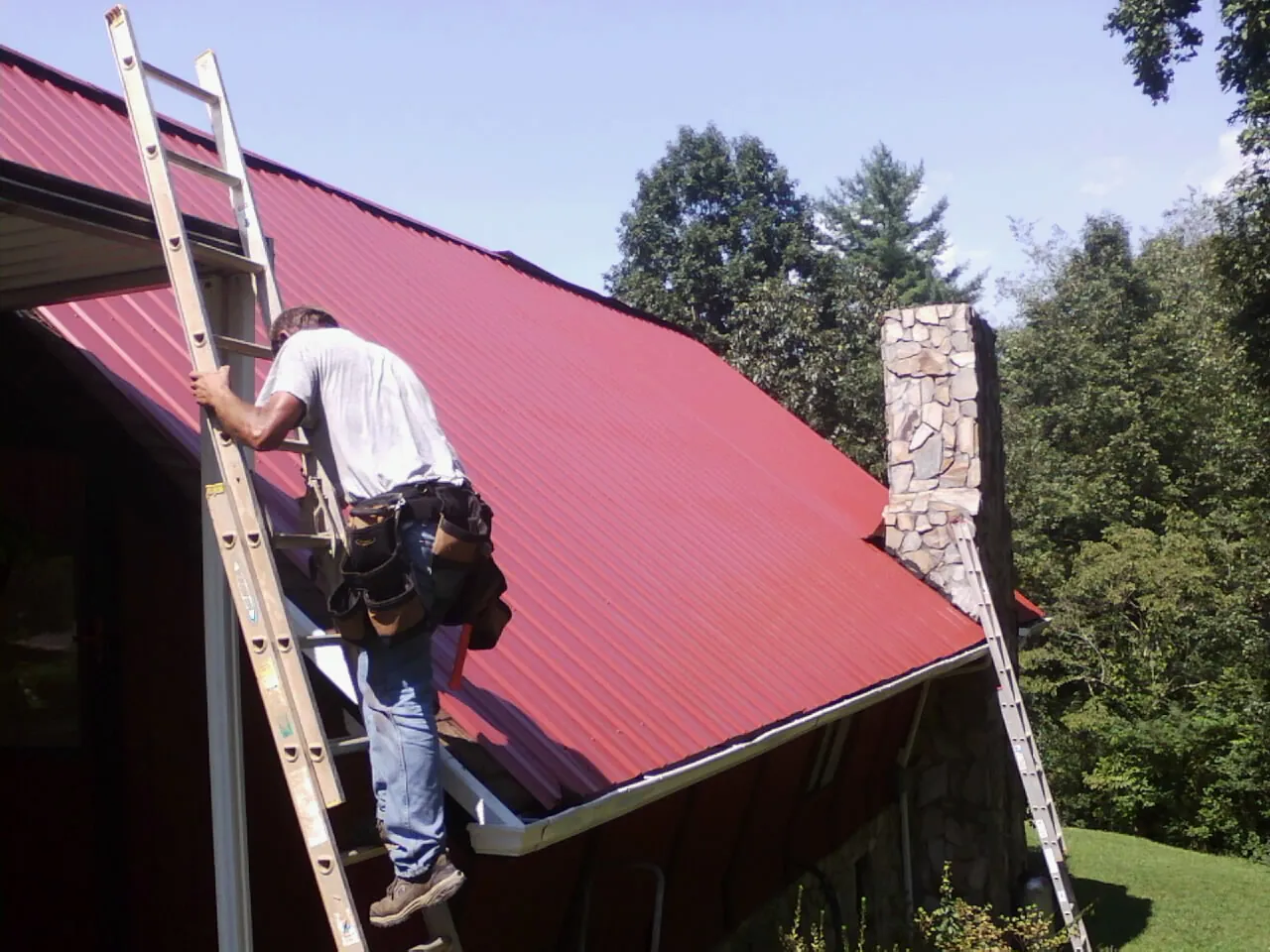 Roofer on a ladder beside a red metal roof and stone chimney — Soffit & Fascia Repair by Roofing Brothers in Neelyton